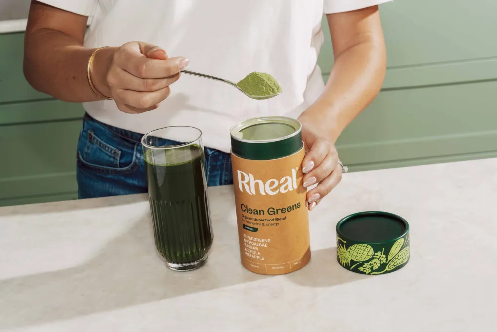 A person spoons Rheal Balance Tonic powder into a glass of water next to the product container.