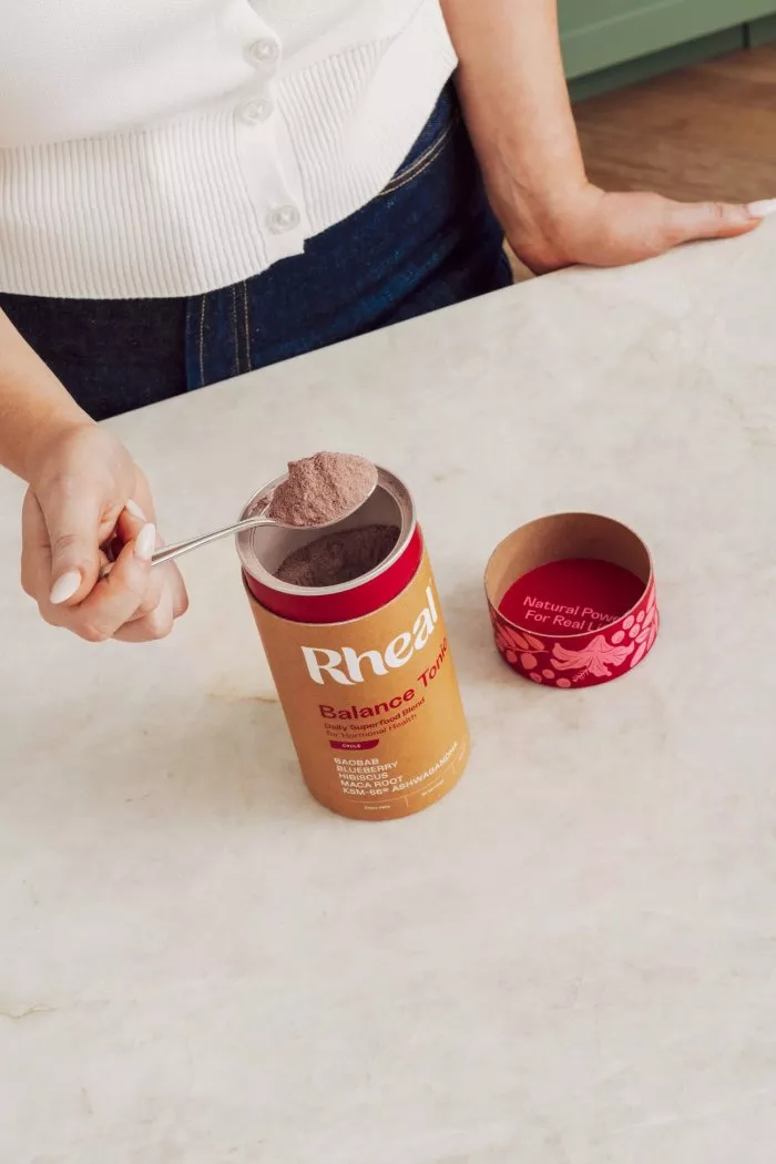 A person spoons a pinkish-brown powder from a container of Rheal Balance Tonic onto a marble countertop.