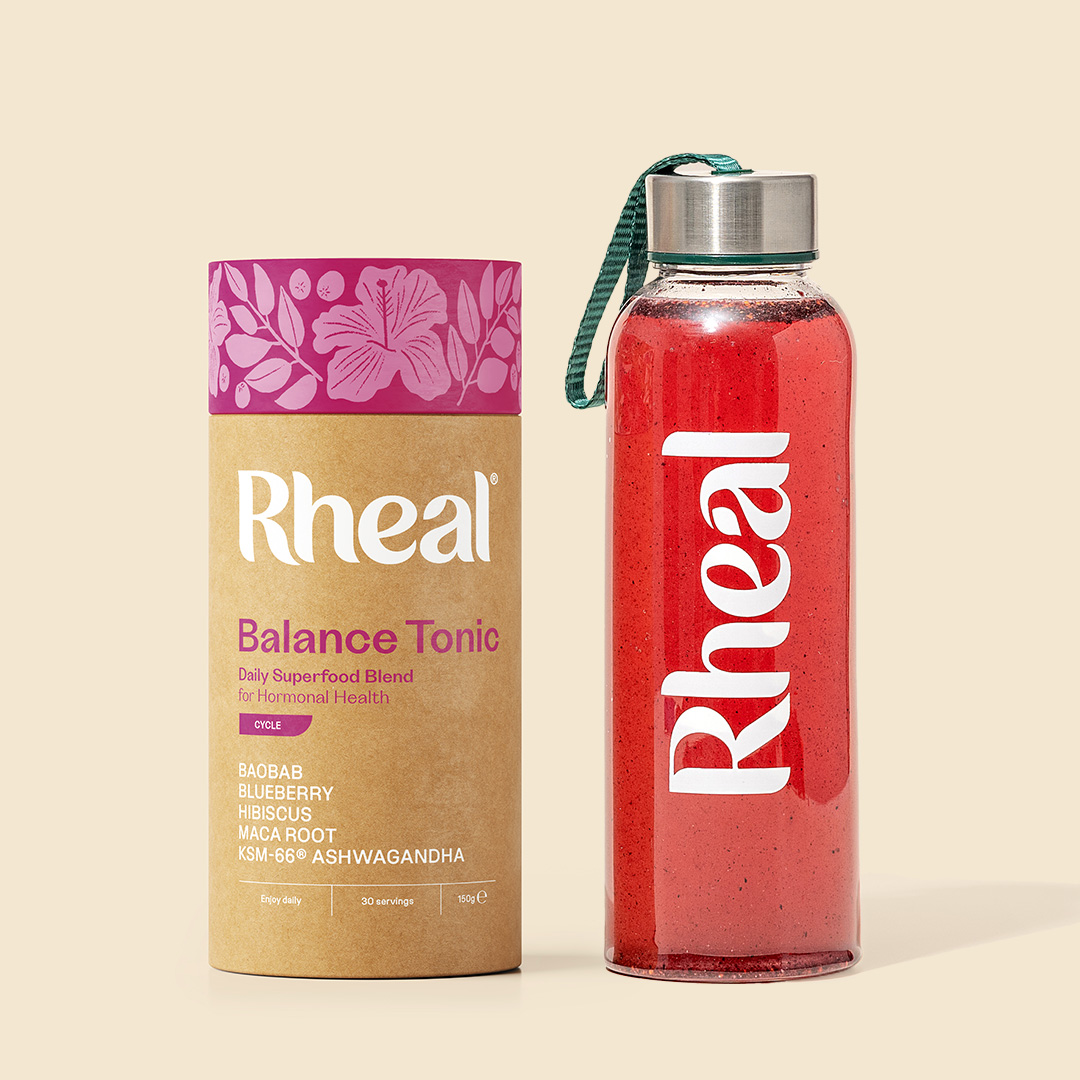 A tube of Rheal Balance Tonic next to a glass bottle containing a red beverage.
