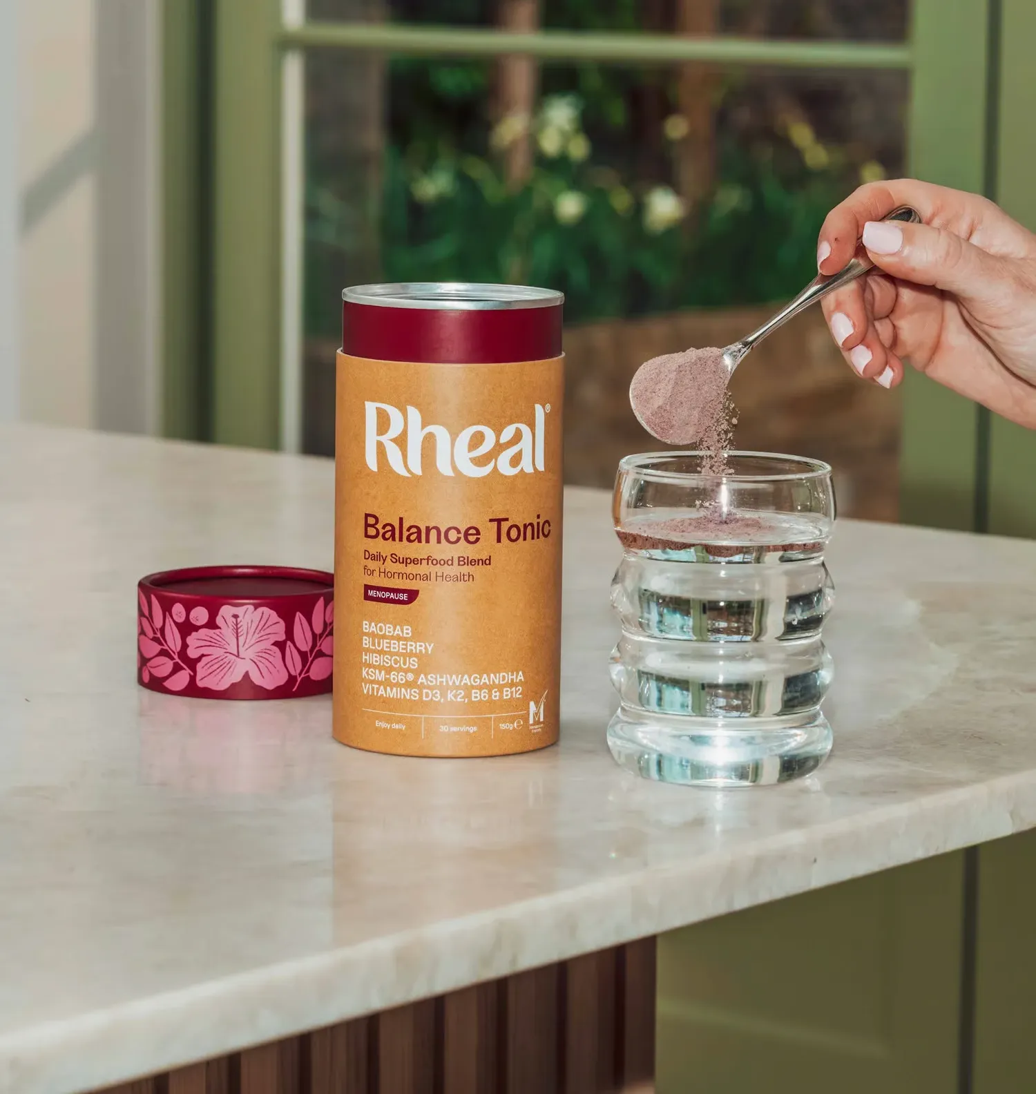 A person spoons Rheal Balance Tonic powder into a glass of water next to the product container.