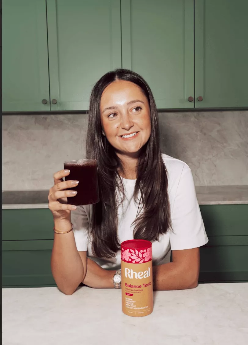 A smiling woman in a white t-shirt holds a dark beverage next to a container of Rheal Balance Tonic.
