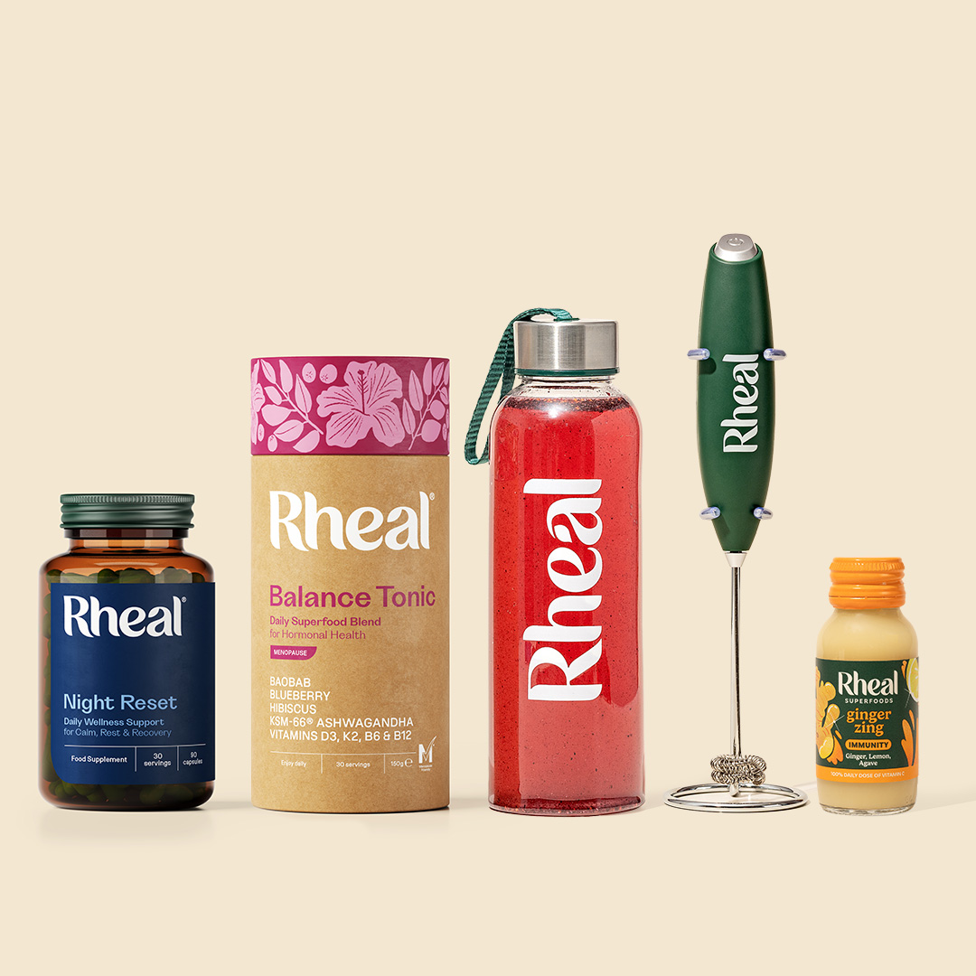 A lineup of Rheal brand wellness products including capsules, powders, a drink bottle, a whisk, and a shot.