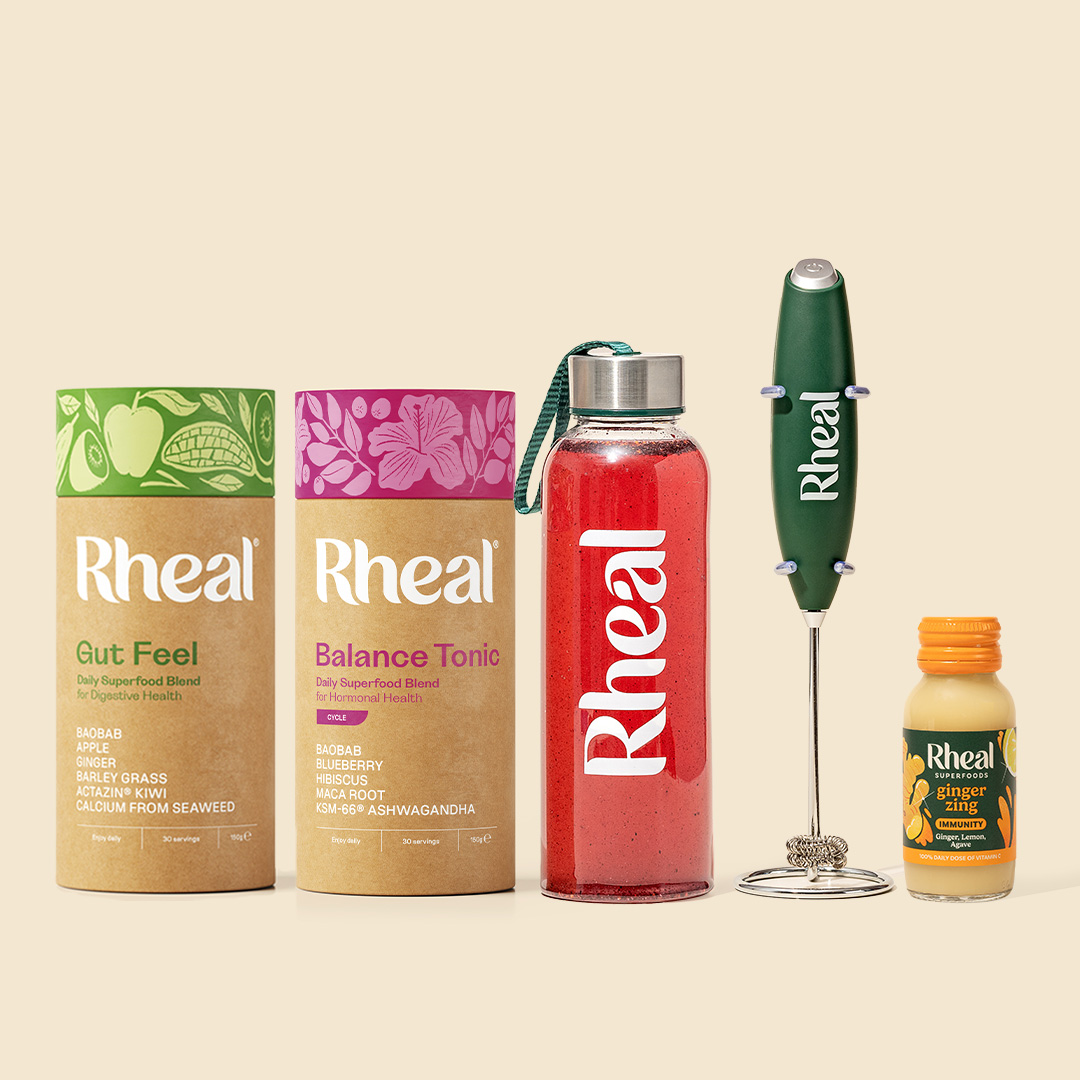 A collection of Rheal superfood products including powders, a drink bottle, a whisk, and a small shot.