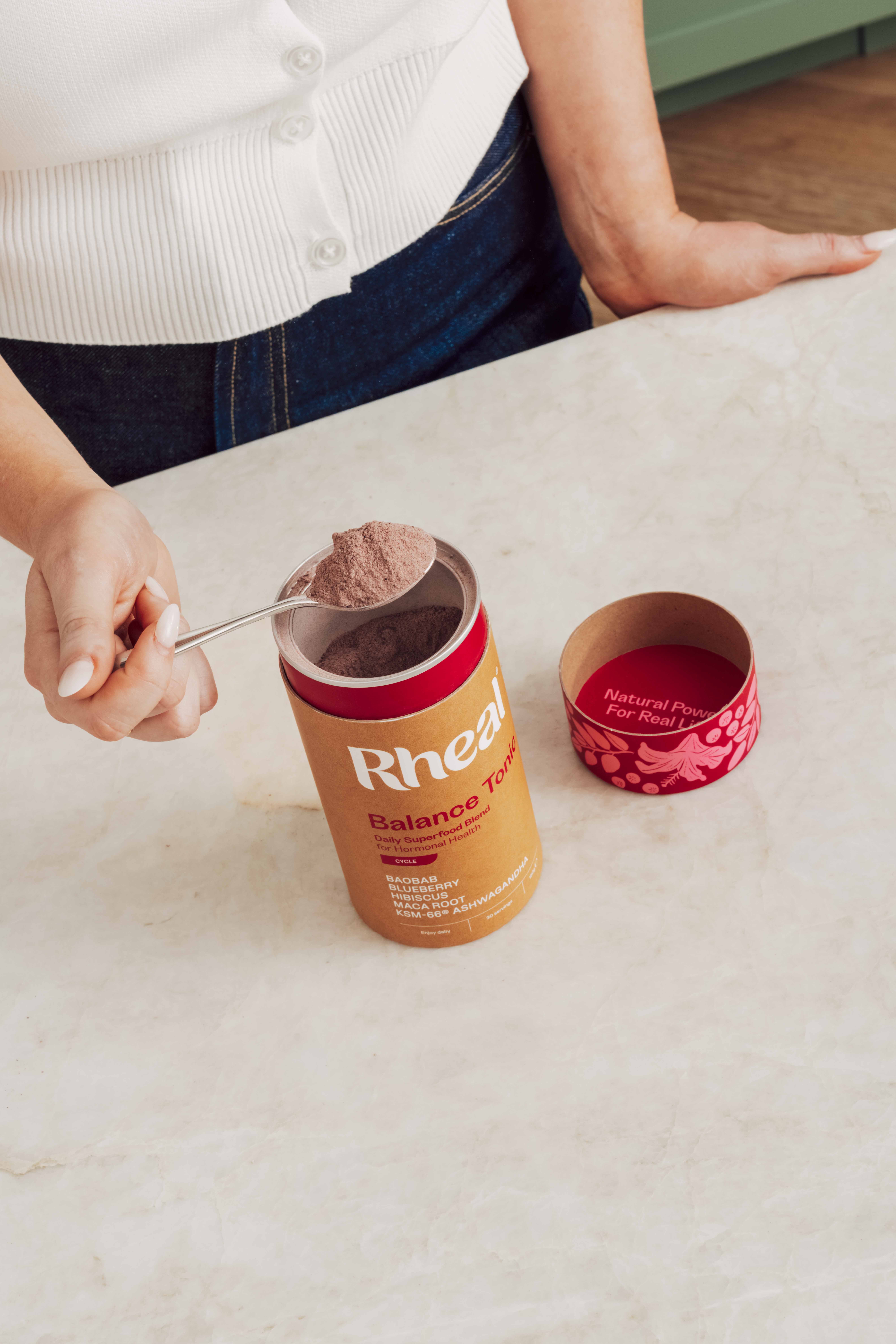A person scoops a spoonful of Rheal Balance Tonic powder from its container on a marble countertop.