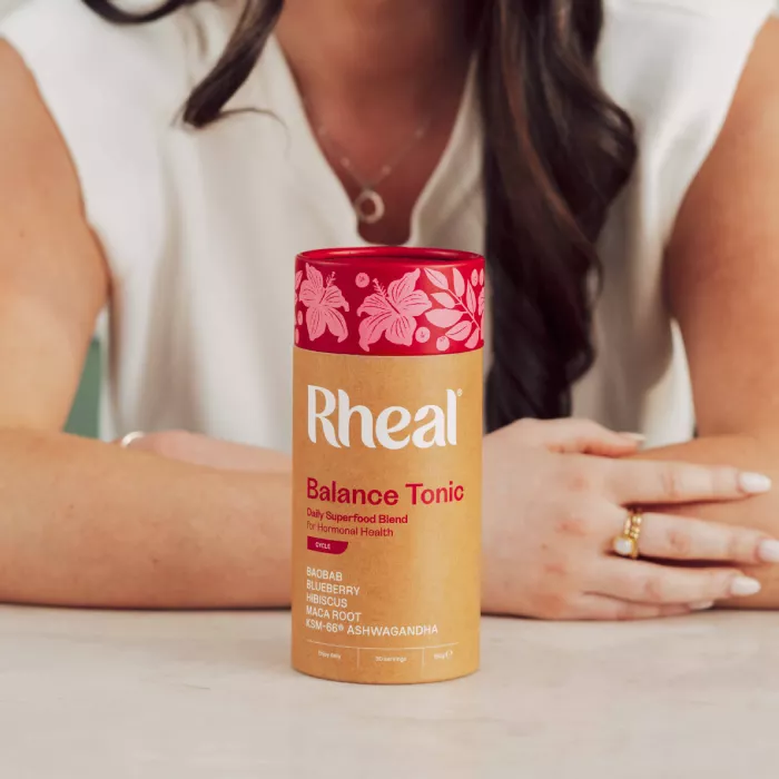 A woman sits at a table with a container of Rheal Balance Tonic superfood blend in front of her.