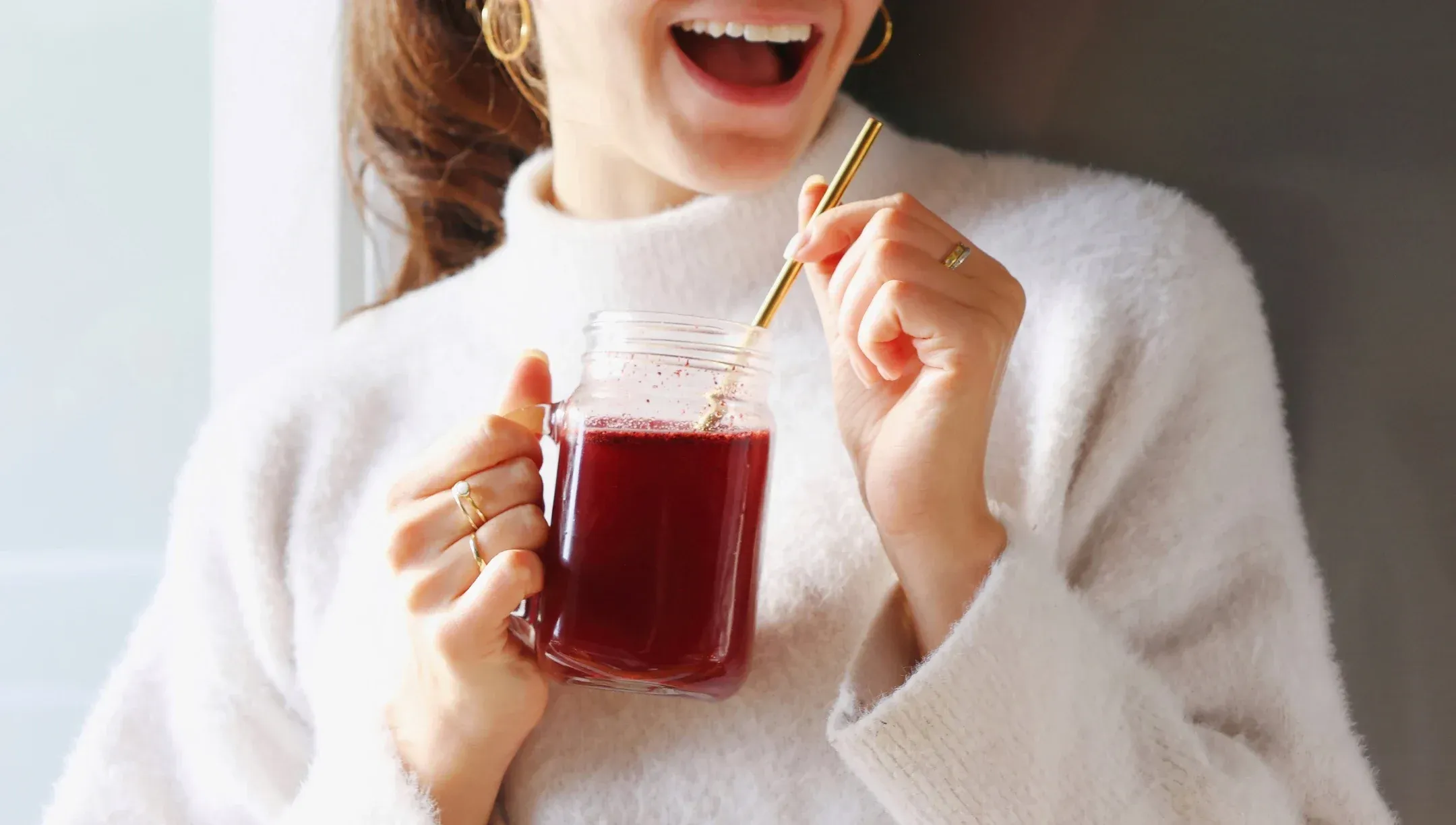 A smiling woman in a white sweater holds a red drink in a mason jar with a straw.