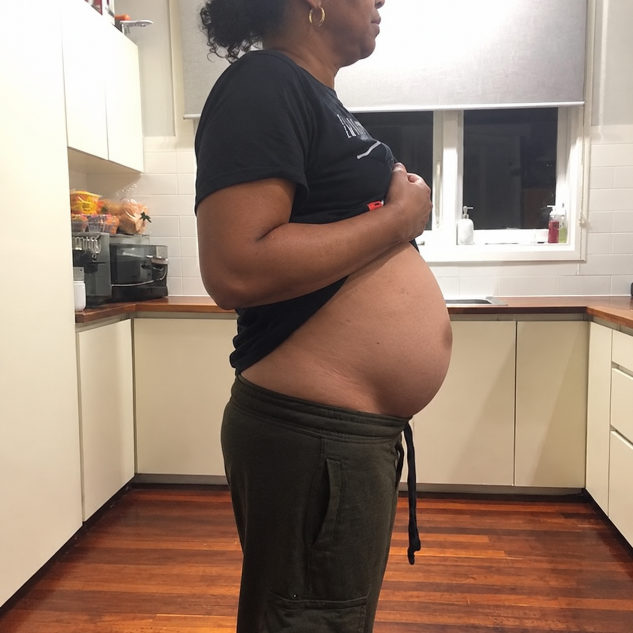 A person stands in profile in a kitchen, holding their shirt up to show their pregnant belly.