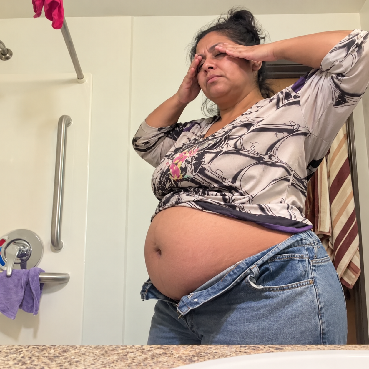 A woman in a bathroom with her hands on her forehead, her shirt lifted over her stomach.