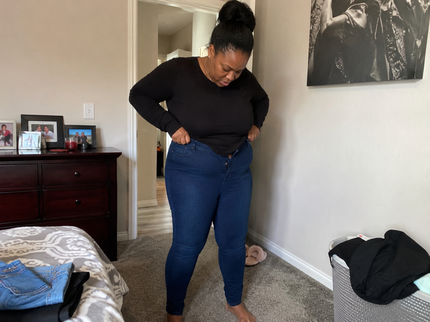 A woman in a black shirt stands in a bedroom, pulling on a pair of blue jeans.