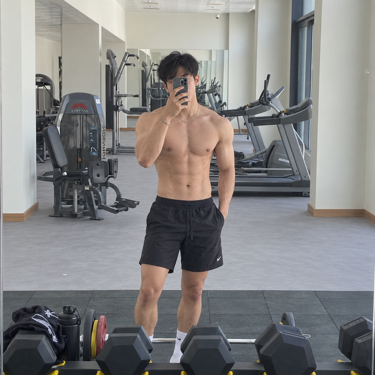 A shirtless, muscular man takes a selfie in a gym mirror, surrounded by workout equipment.