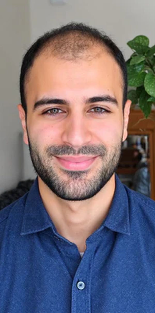 A headshot of a man with short dark hair and a beard, smiling in a blue collared shirt.