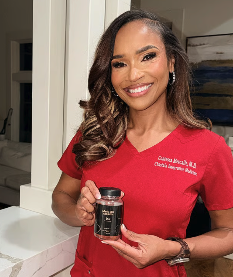 A smiling woman in red scrubs holds up a bottle of Black Girl Vitamins D3.