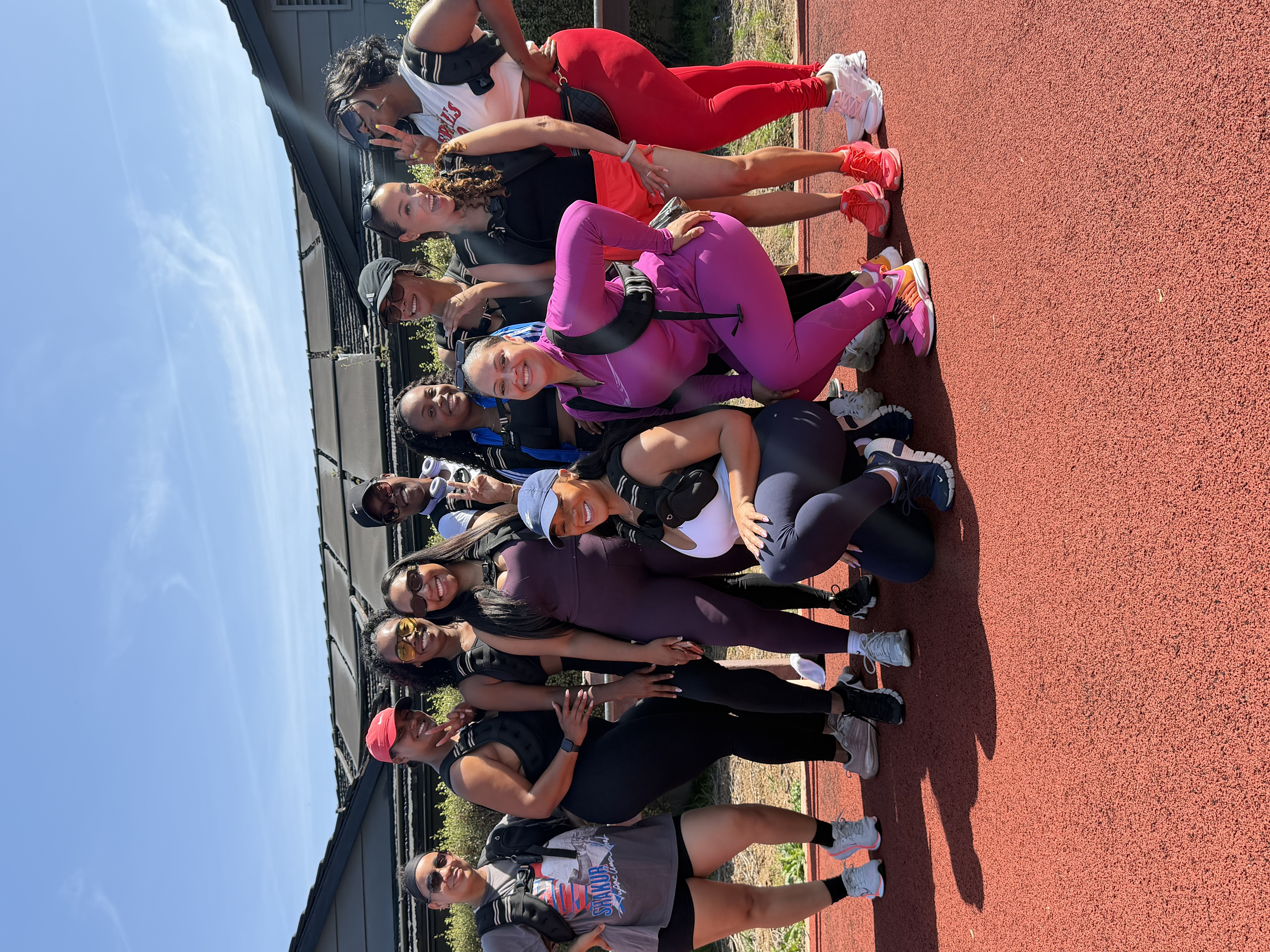 A group of eleven women in athletic gear posing for a photo on a red track outdoors.