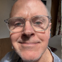 A close-up photo of a smiling bald man wearing clear-framed glasses.