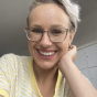 A close-up of a smiling woman with short blonde hair and glasses, resting her head on her hand.