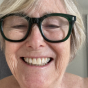 A close-up of an older woman with short blonde hair and dark-rimmed glasses, smiling with her eyes closed.