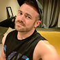 A man with a beard, wearing a backwards baseball cap and a black sleeveless shirt, looks at the camera.