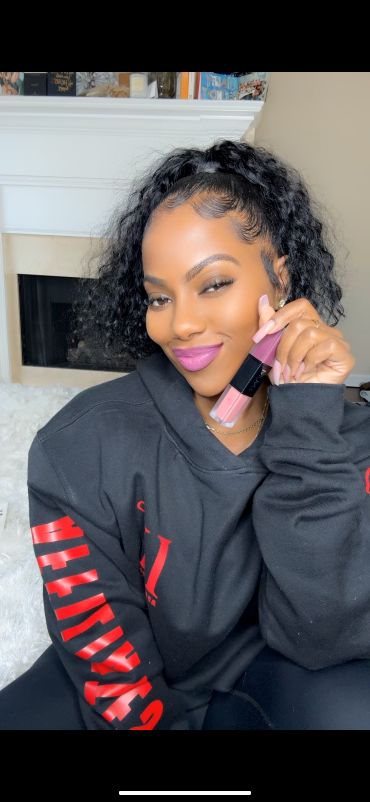 Person smiling, holding a pink lipstick tube, wearing a black hoodie.