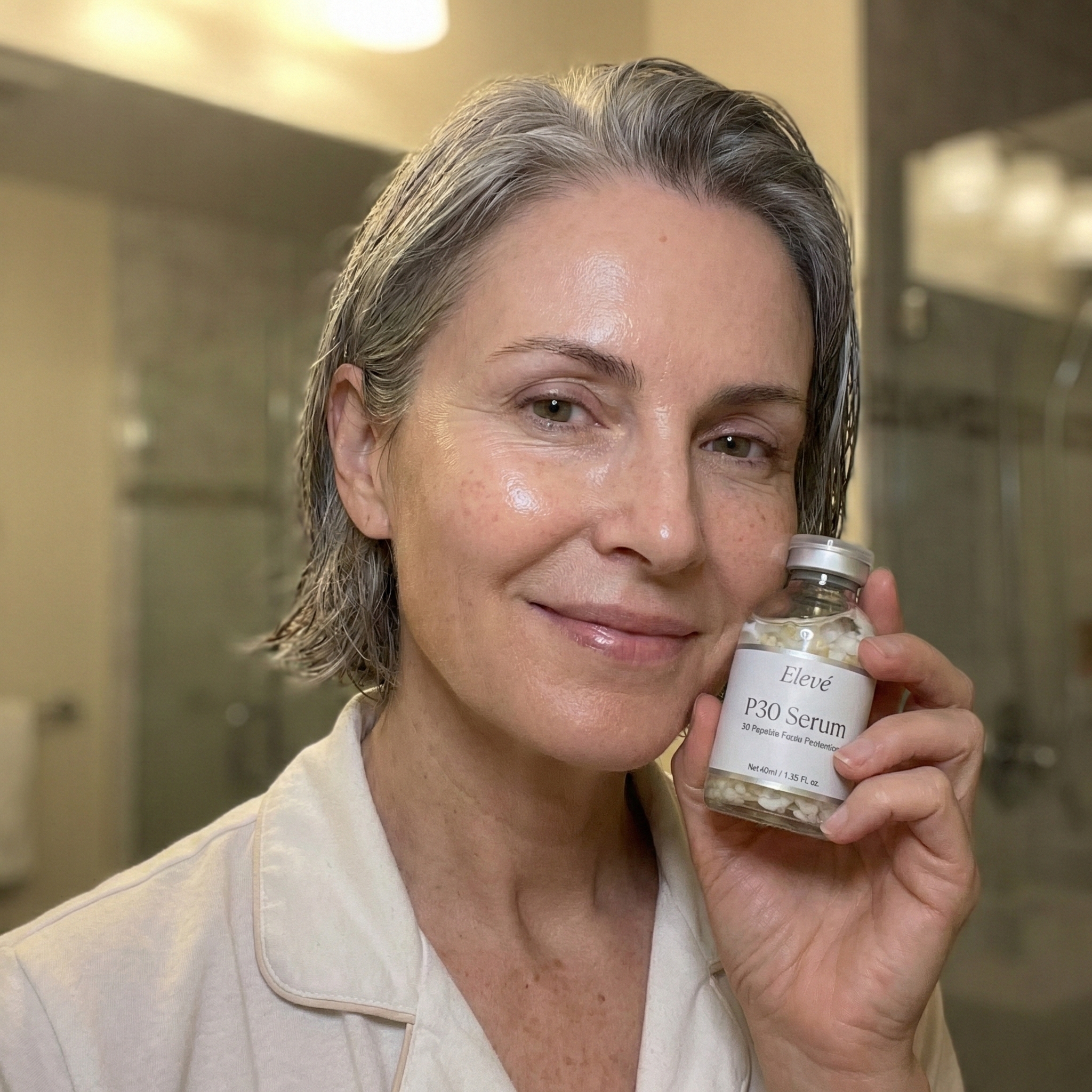 A smiling woman with gray hair holds a small glass bottle of Elevé P30 Serum.