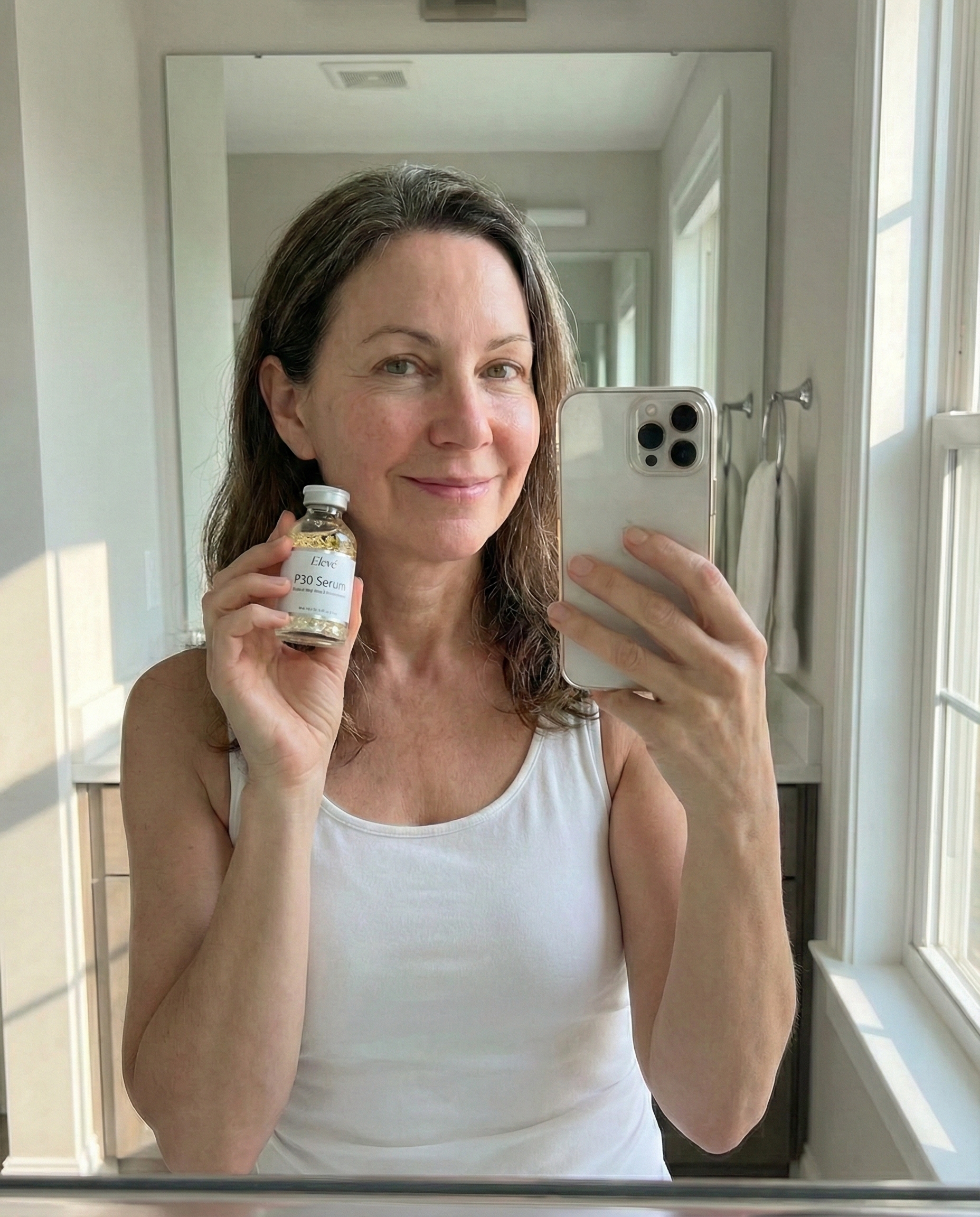 A woman takes a mirror selfie in a bathroom, holding a phone and a bottle of serum.
