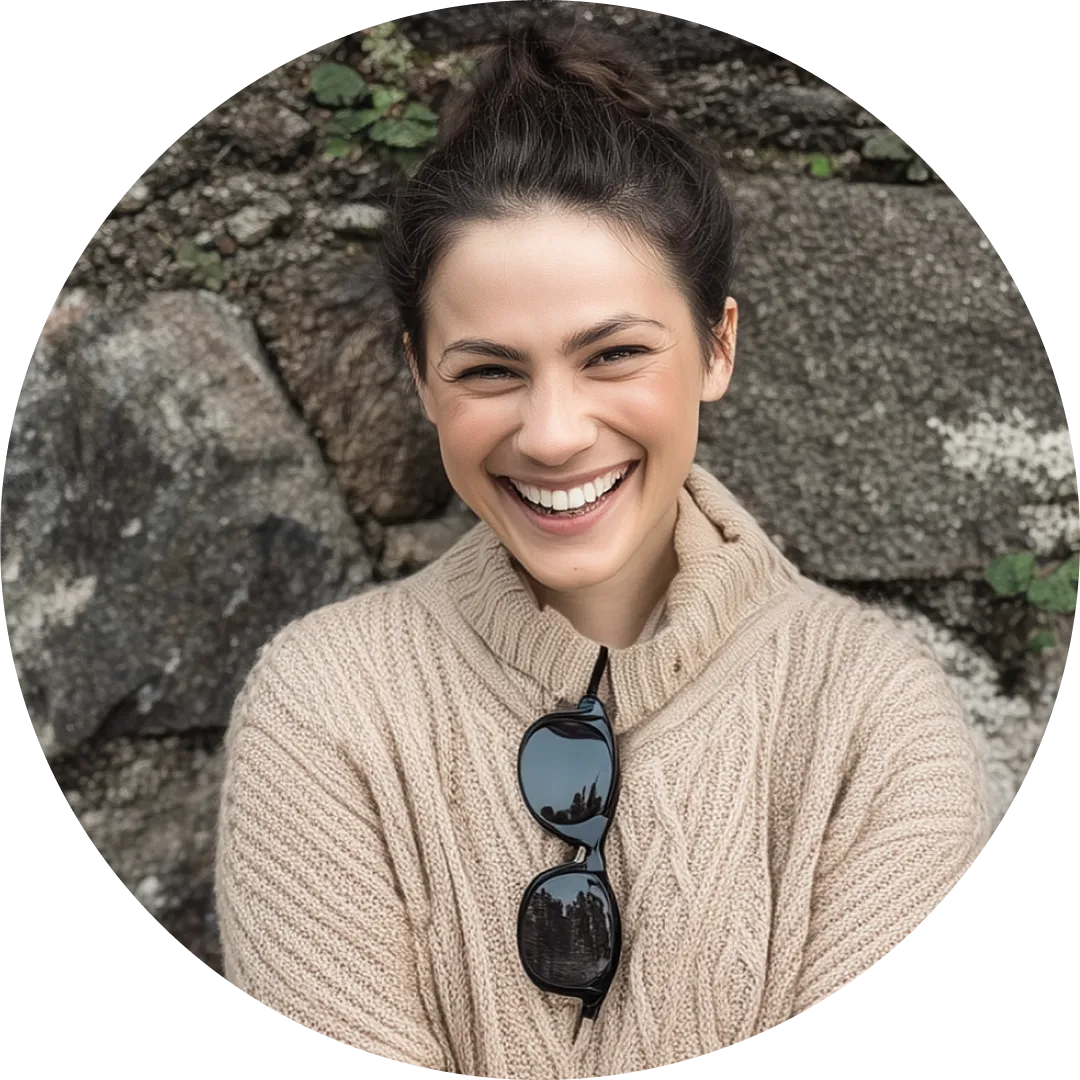 Smiling person in a beige sweater with sunglasses, standing against a stone wall.
