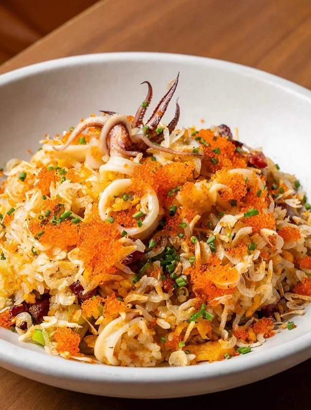 A close-up of a white bowl filled with seafood fried rice, topped with squid and orange roe.