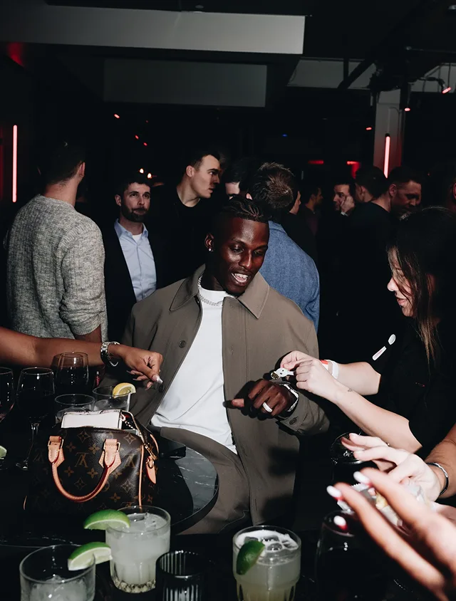 A smiling man sits at a table with people and drinks in a dimly lit, crowded club.