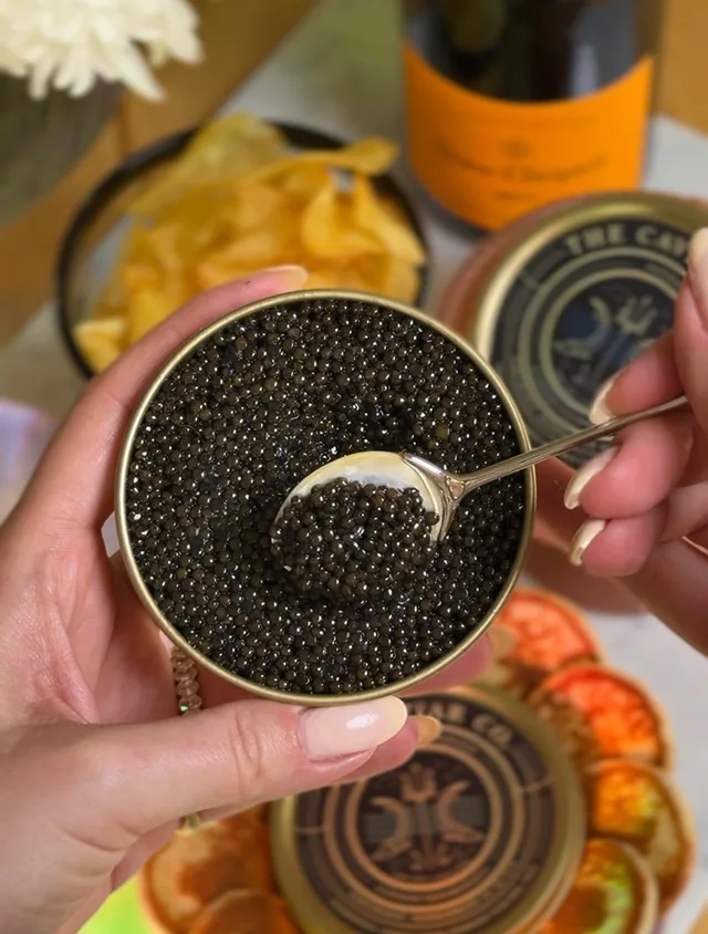 A person holds an open tin of black caviar, scooping some with a small spoon.