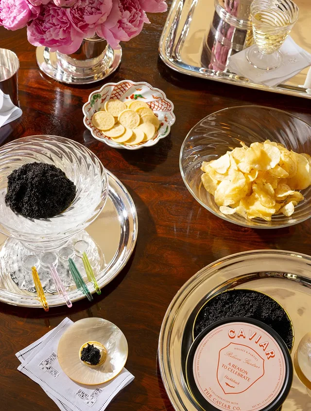 A wooden table set with caviar, crackers, potato chips, silver trays, and a bouquet of pink peonies.