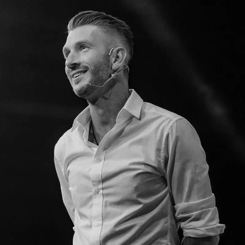 A black and white photo of a smiling man in a white shirt wearing a microphone.