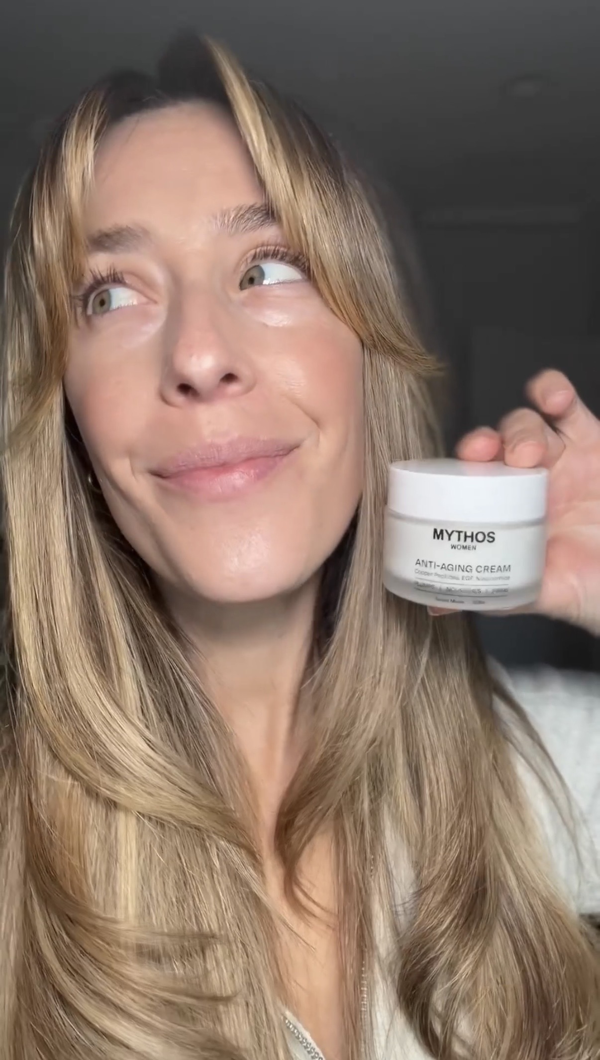 A woman with long blonde hair holds a small white jar of Mythos Anti-Aging Cream.