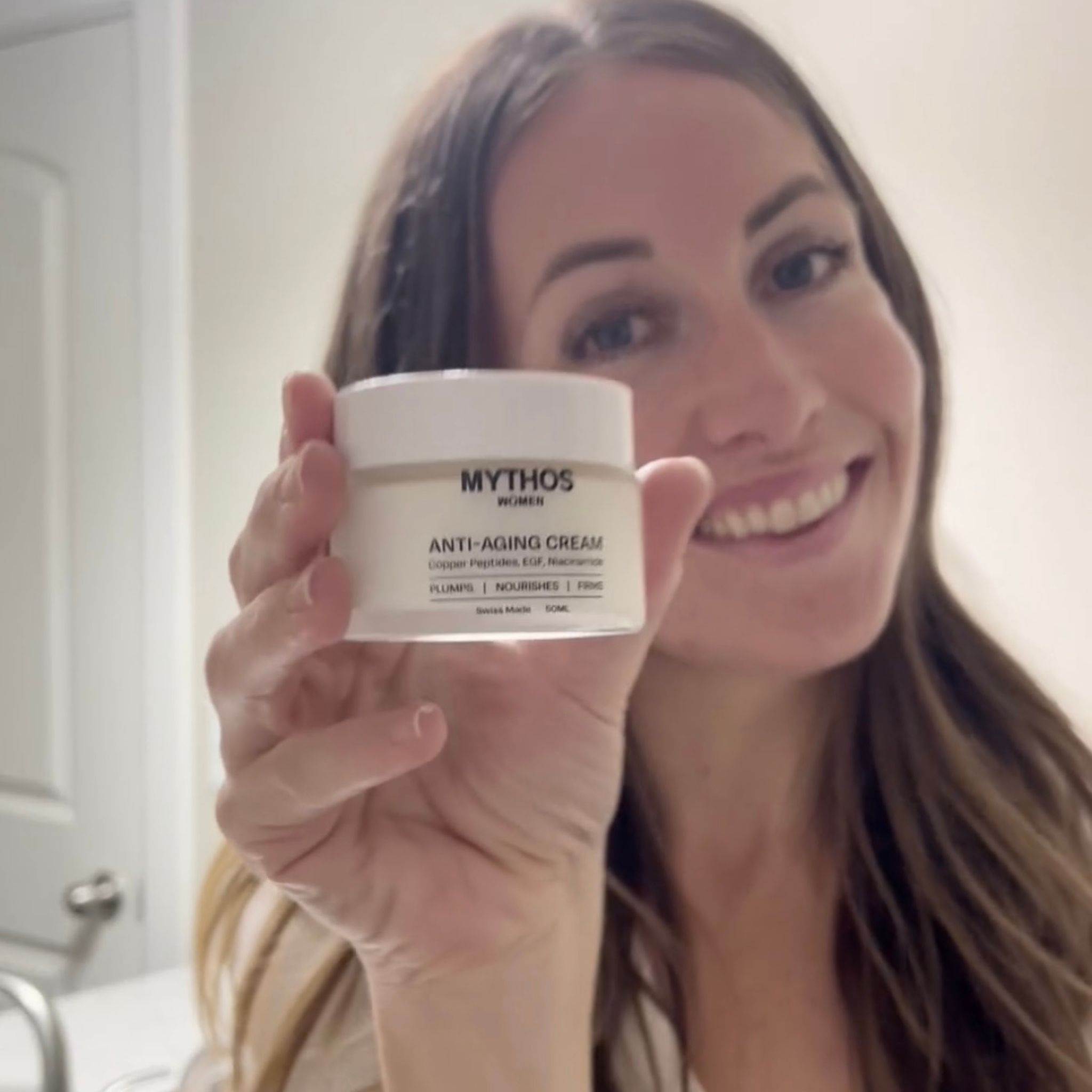 A smiling woman with long brown hair holds up a white jar of Mythos Anti-Aging Cream.