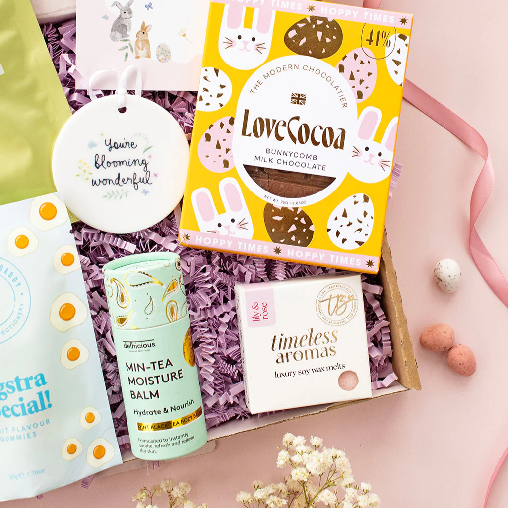 Gift box with chocolate, skincare, wax melts, and decorative items on pink background.