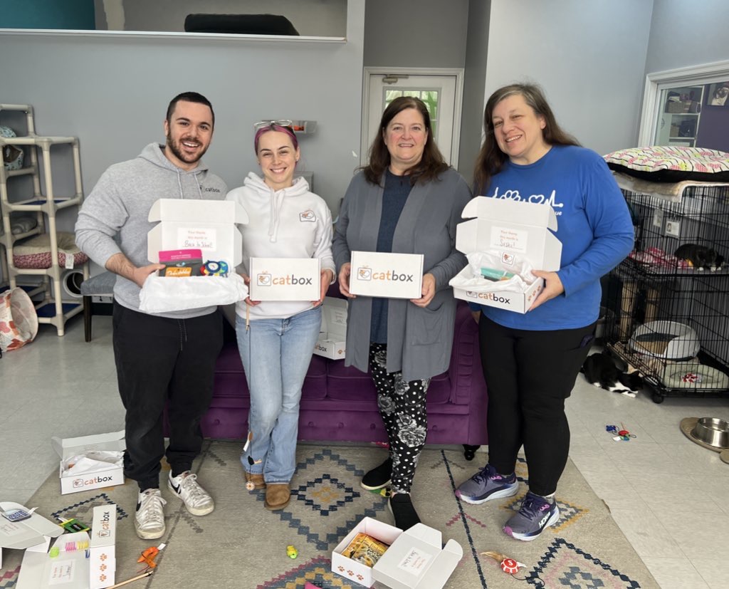 Four people holding 'catbox' packages in a room with cat toys and cages.