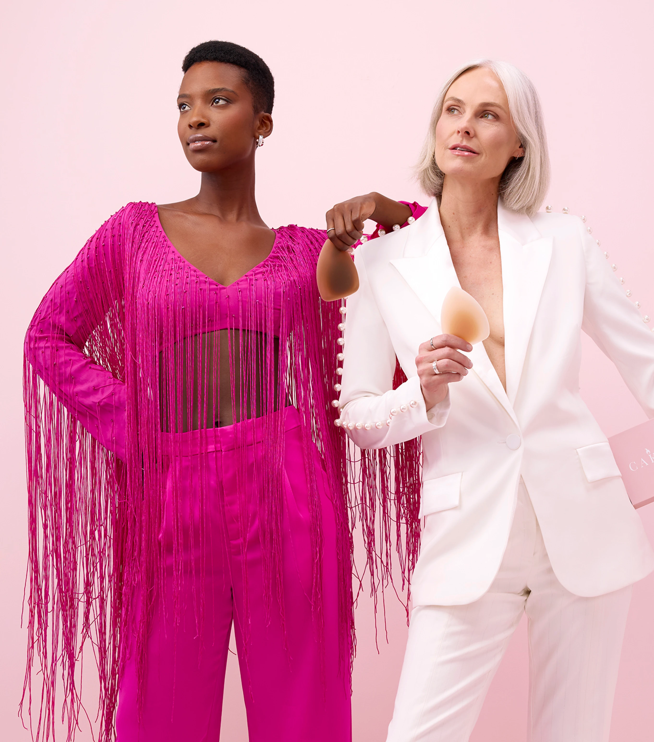 Two women in stylish outfits holding makeup sponges against a pink background.