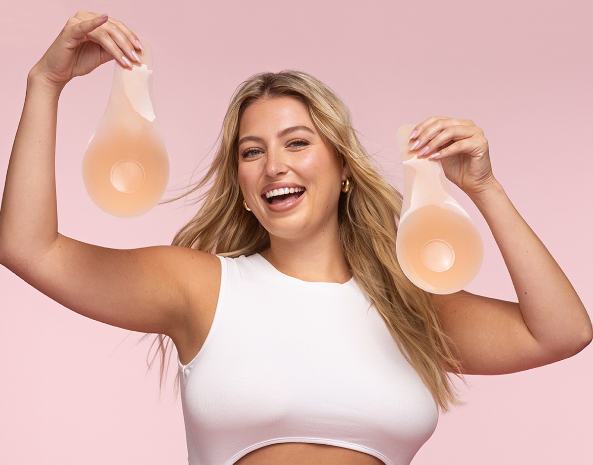 A smiling woman in a white top holds up two adhesive breast lift pasties against a pink background.