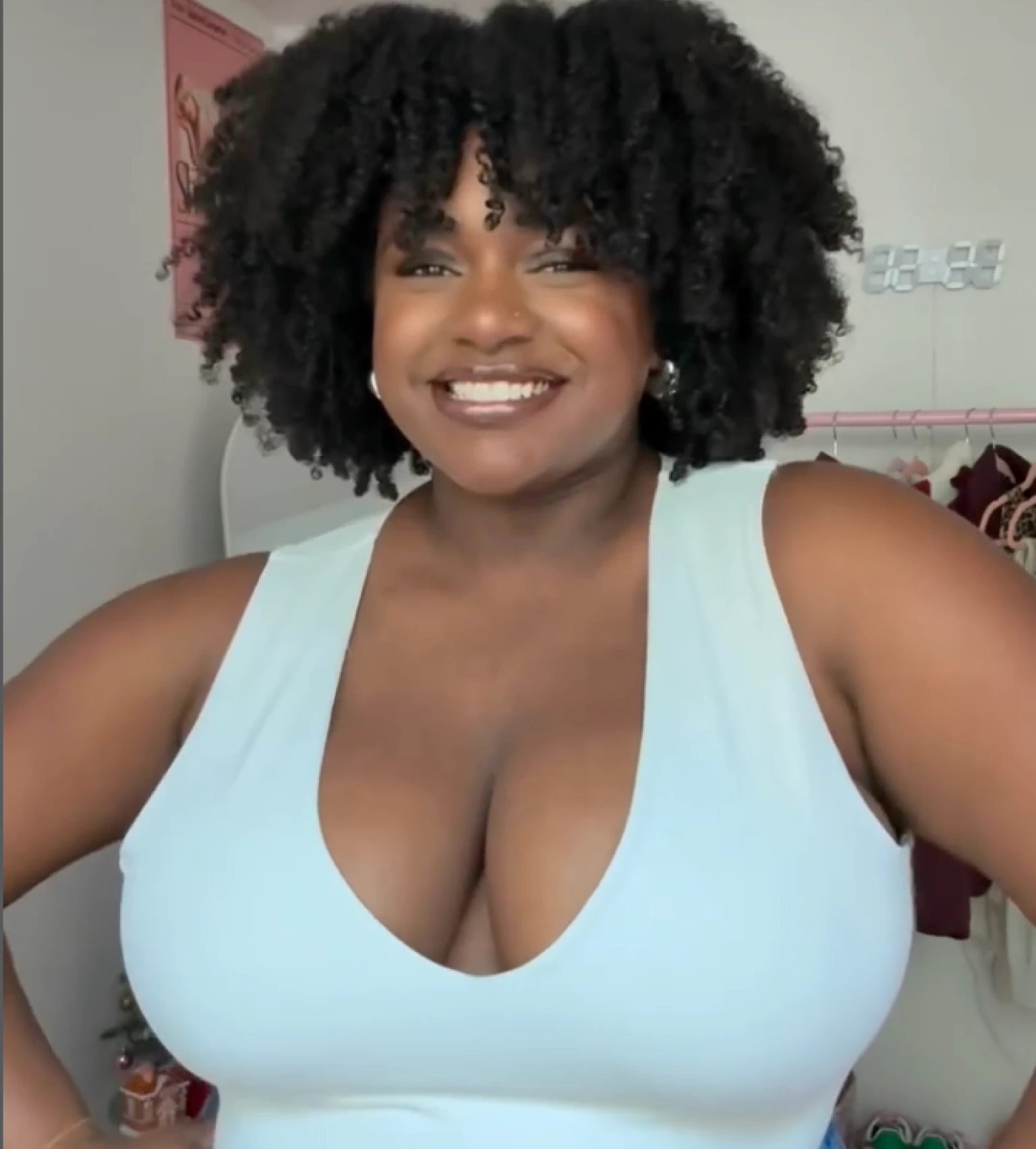 A person smiling, wearing a light blue top, with curly hair in a room setting.