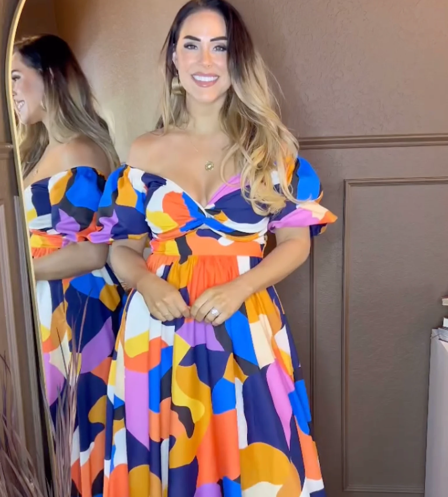 Woman in a colorful dress standing by a mirror, smiling.