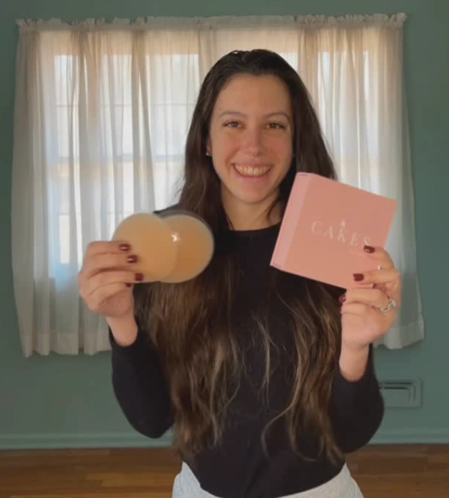 Person holding a pink box and adhesive silicone pads, smiling in front of curtains.