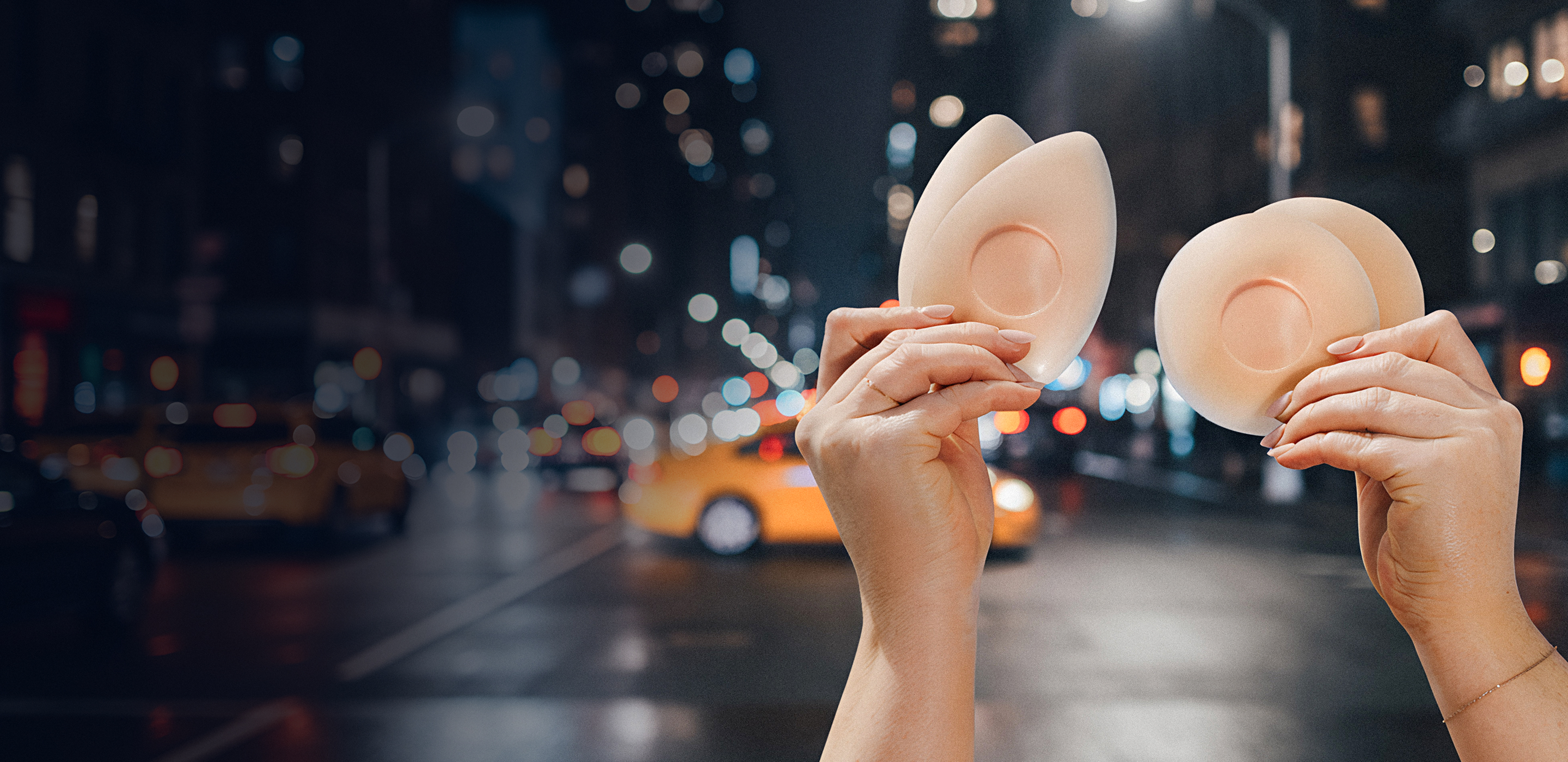 Hands holding bra inserts against a city street background at night.