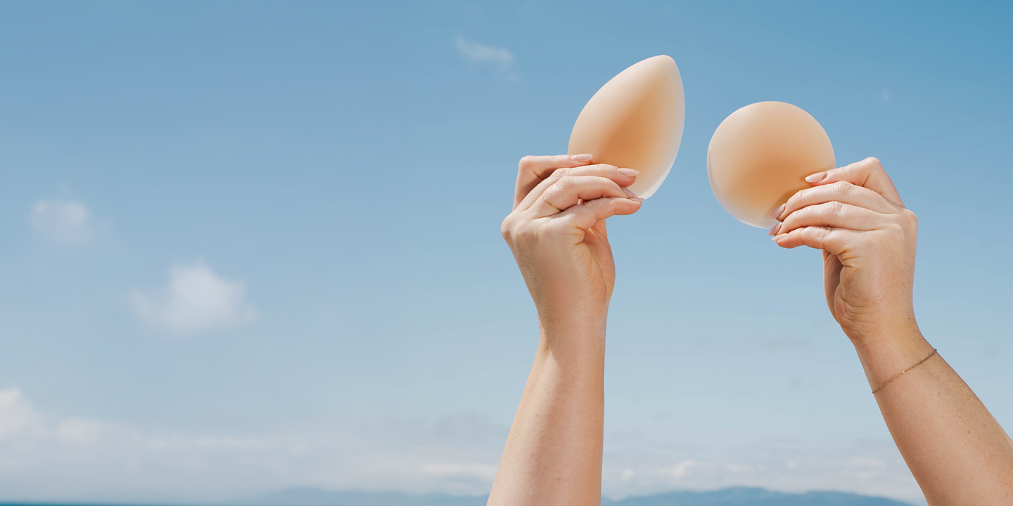 A pair of hands hold two different types of breast implants up against a blue sky.