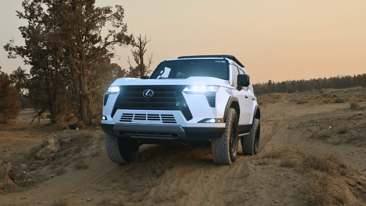 A white Lexus SUV drives on a dirt road through a dry landscape with scattered trees.