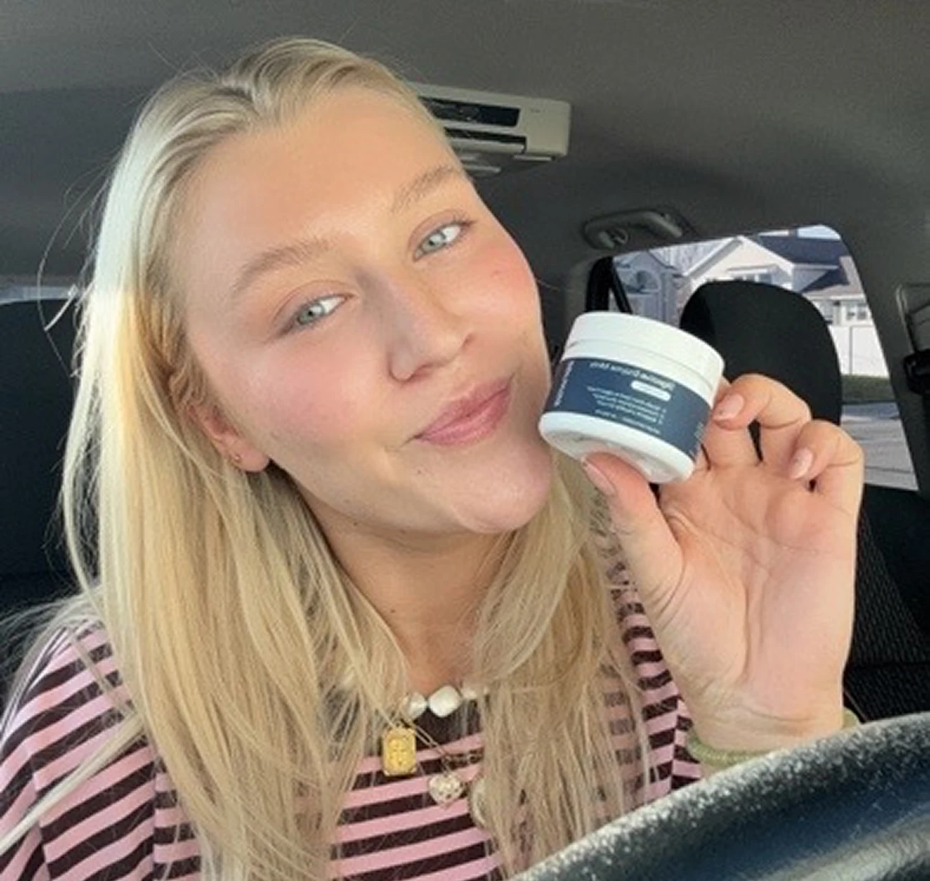 A blonde woman in a striped shirt sits in a car, smiling and holding a small white jar.