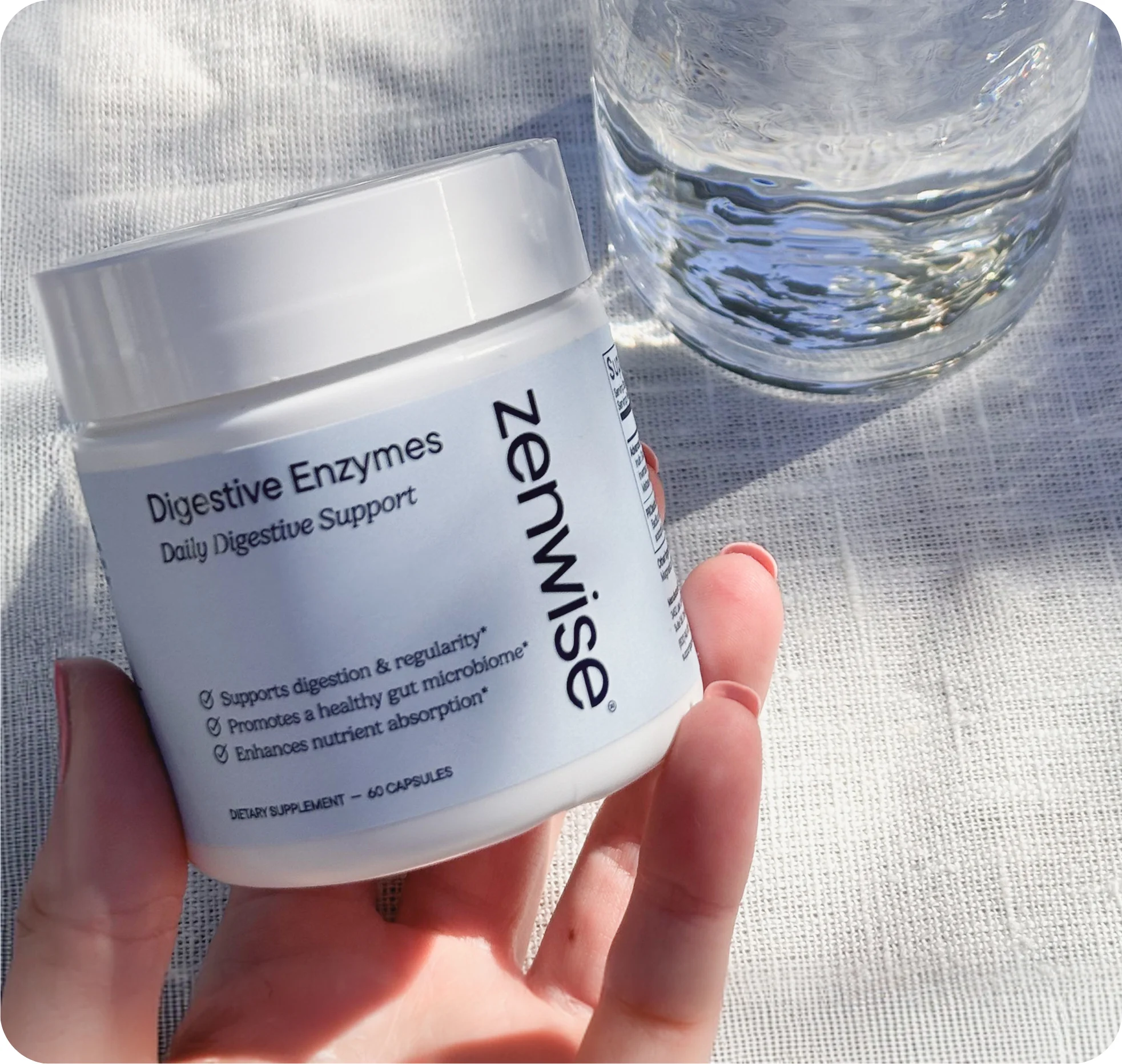 A hand holds a white bottle of Zenwise Digestive Enzymes next to a glass of water.