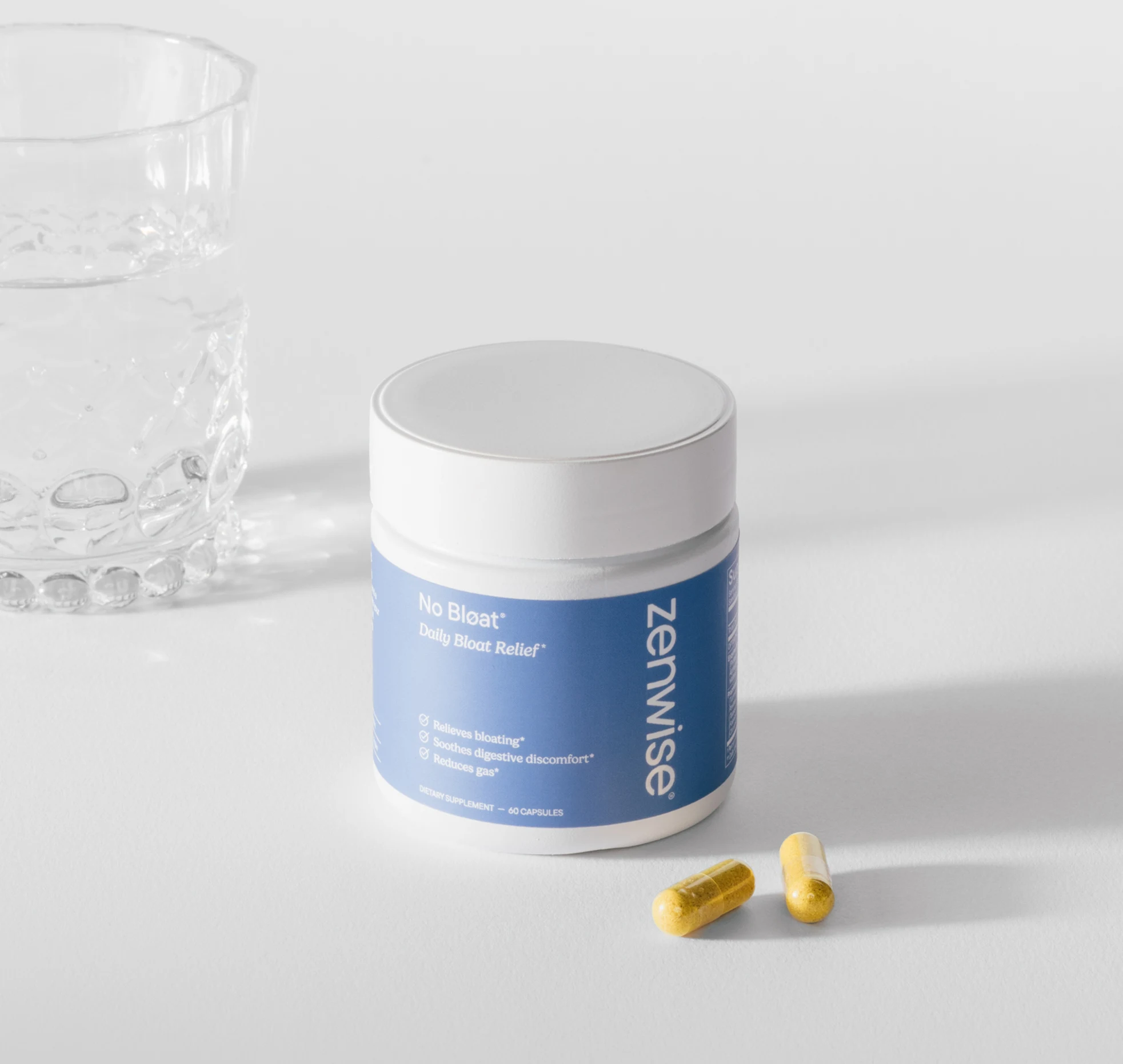A bottle of Zenwise No Bloat supplement with two capsules and a glass of water on a white surface.