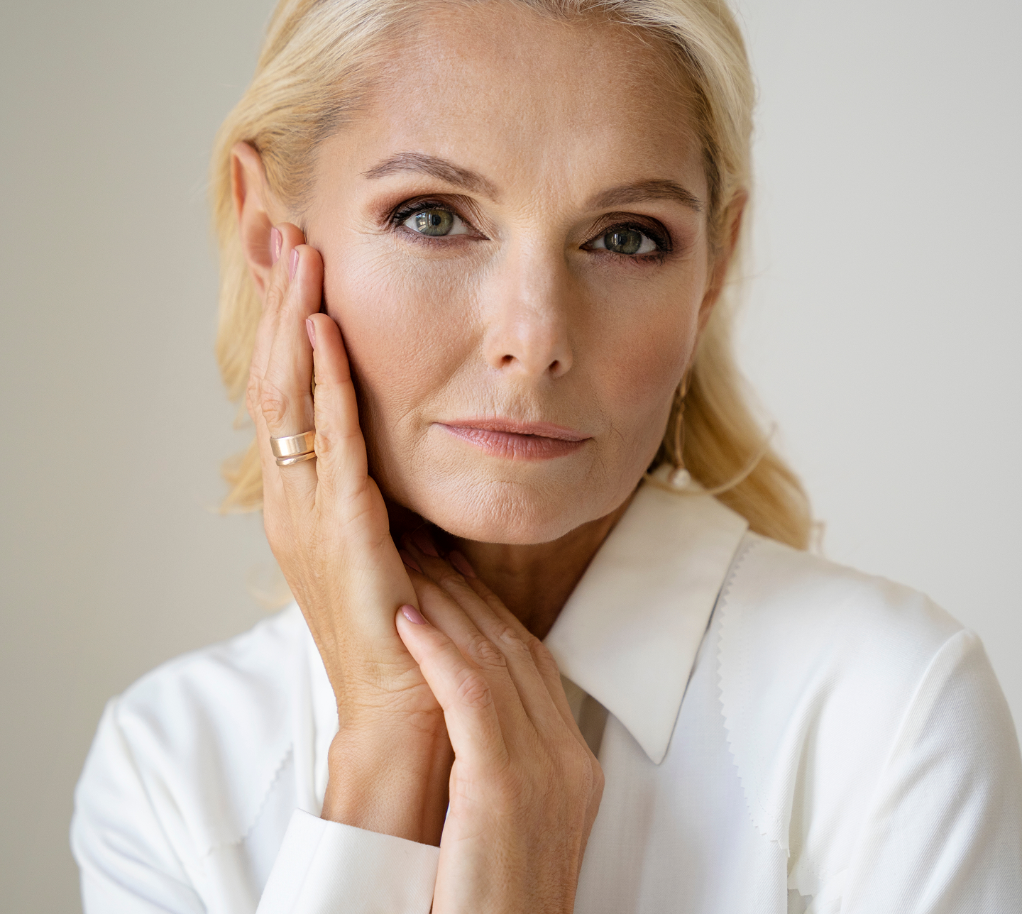 A middle-aged blonde woman in a white shirt looks at the camera, with her hands on her cheek and neck.