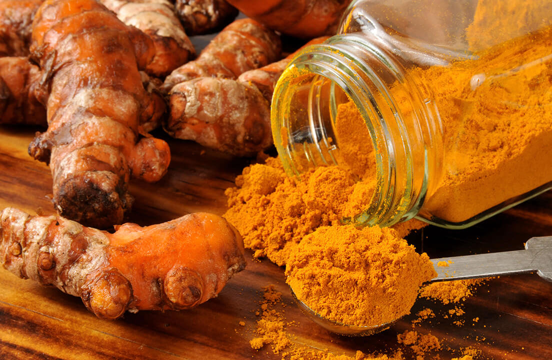 Turmeric roots and spilled turmeric powder from a glass jar on a wooden surface.
