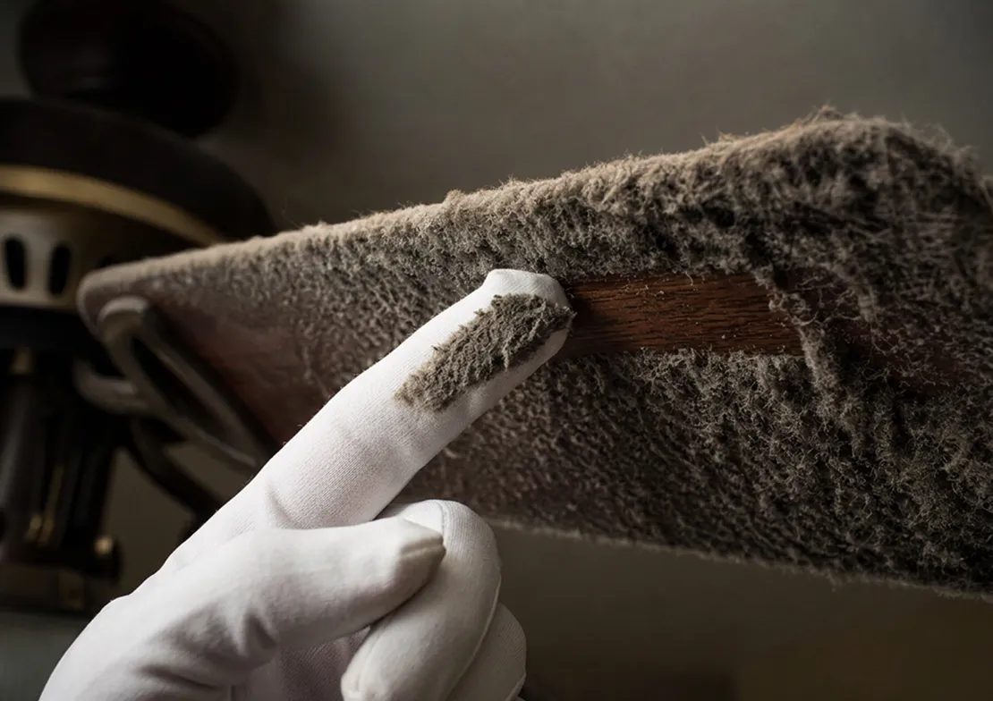 A white-gloved finger wiping a thick layer of dust from a ceiling fan blade.