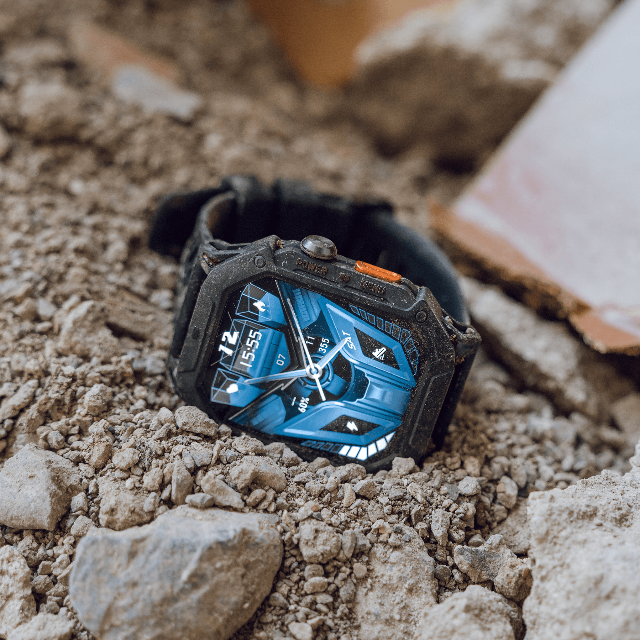 A rugged smartwatch with a blue display on rocky ground.