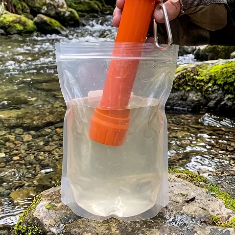 A hand places an orange portable water filter into a clear pouch of water by a stream.